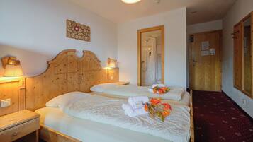 Une chambre d’hôtel avec deux lits en bois, une tête de lit en bois, une table de chevet en bois, une lampe fixée au mur et une porte en bois.