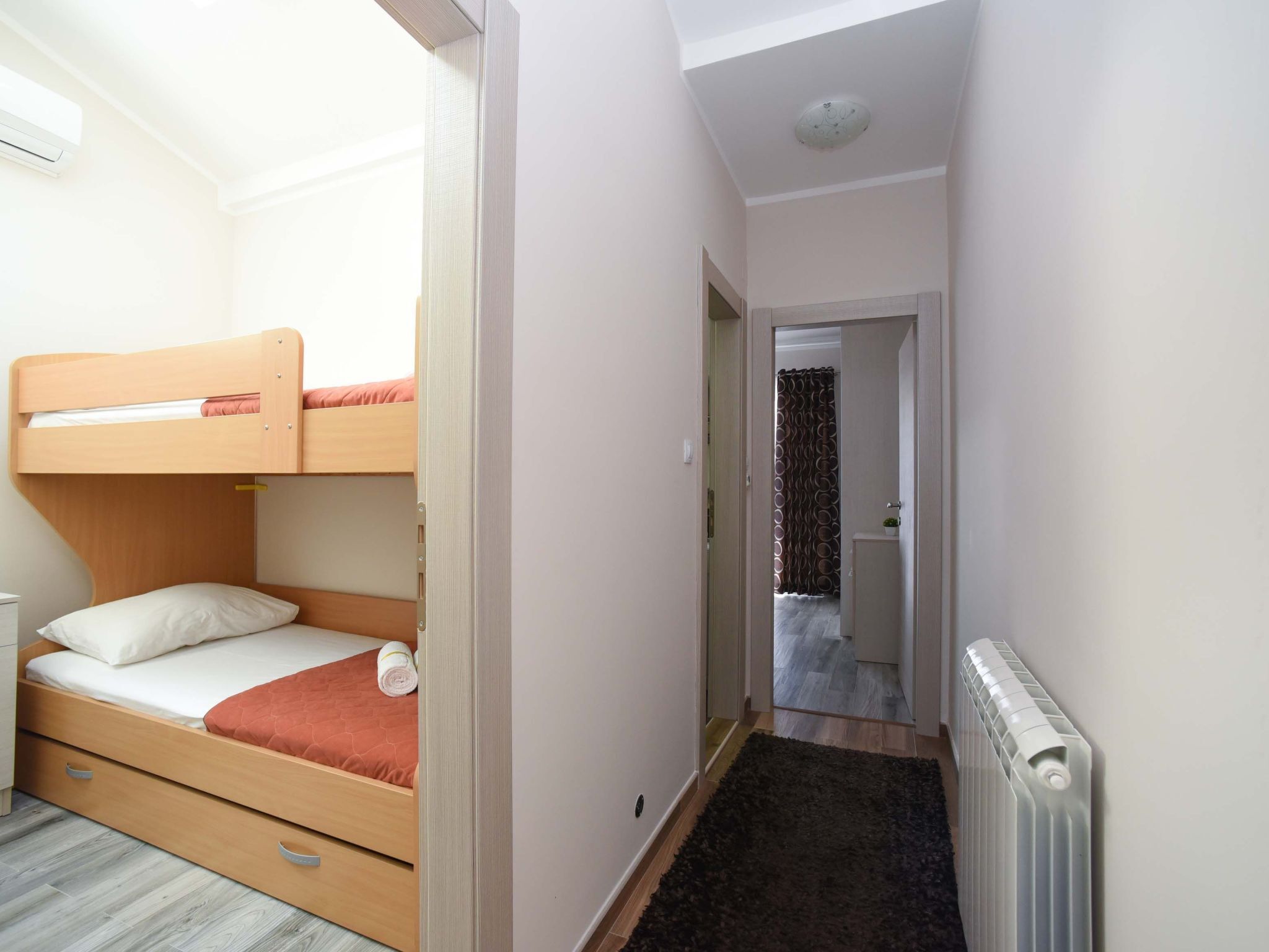 Une petite chambre comprenant un lit superposé, une petite table de chevet, un radiateur et un étroit couloir menant à la salle de bain.