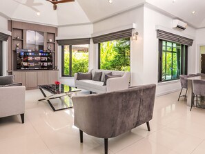 Una sala de estar moderna con un sofá, una mesa de centro y un televisor. Hay ventanas grandes con cortinas, un aire acondicionado y un ventilador de techo.