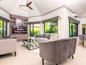 A modern living room with a sofa, armchairs, a coffee table, and a TV. There is a kitchen area with a refrigerator and a dining table. Large windows provide views of greenery outside. - "Villa Sabai" mit Terrasse by Interhome (Pattaya)