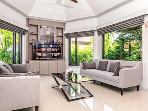 Una sala de estar moderna con sofá, mesa de centro, mueble para televisión y ventanas amplias con vista a zonas verdes.