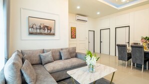 A modern living room with a grey sectional sofa, a marble coffee table, and a painting of horses. There is a dining area with a table set for four and a chandelier.