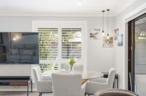 A modern living room with a sofa, a dining table with chairs, a TV, and a window with blinds.