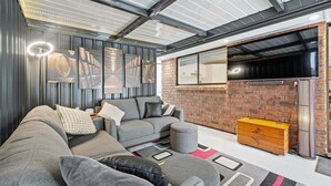 A modern living room with a grey sofa, a coffee table, and a brick accent wall. - Belle Escapes - Fulton Retreat (Holdfast Bay)