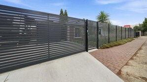 A modern fence with horizontal slats, a paved walkway, and a clear sky. - Belle Escapes - Fulton Retreat (Holdfast Bay)