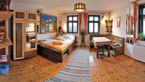 Une pièce confortable avec un lit, un bureau et une chaise. Il y a des fenêtres avec des rideaux, un radiateur et une plante suspendue.