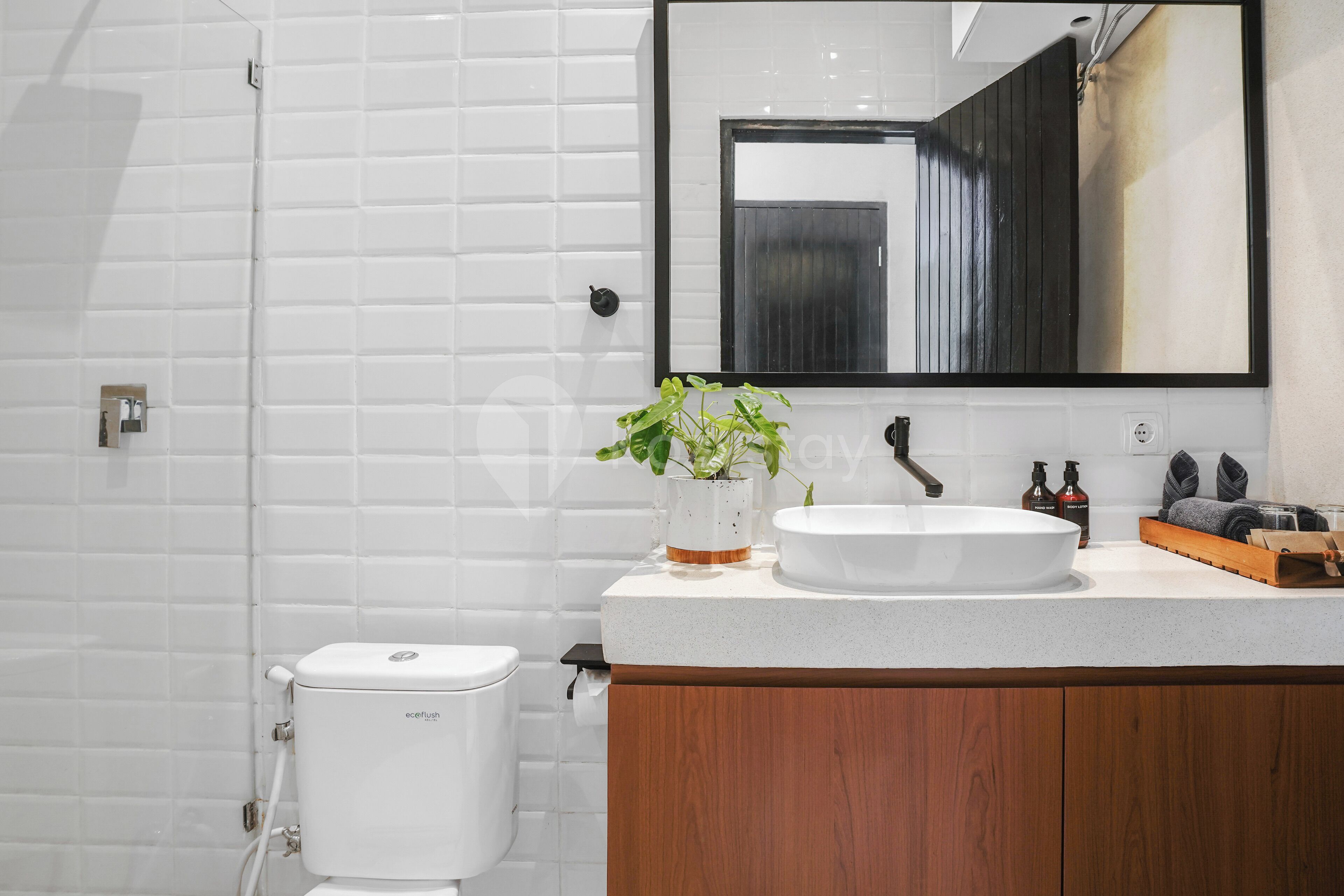 Une salle de bain moderne avec des murs recouverts de carreaux blancs, des toilettes blanches, un meuble-lavabo en bois et un miroir.