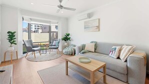 Ein modernes Wohnzimmer mit einem grauen Sofa, einem Holztisch und zwei Sesseln. Es gibt einen Deckenventilator, ein großes Fenster und ein Bild an der Wand.