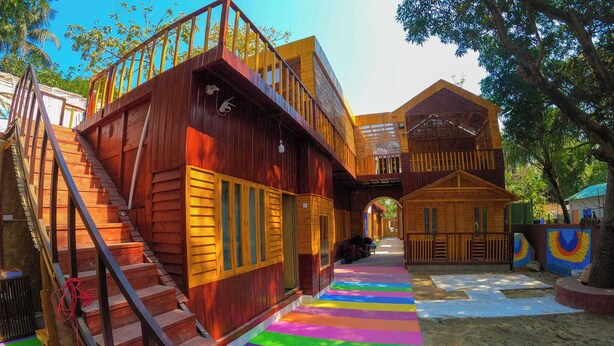 A wooden building with a staircase leading to a balcony, a colorful striped pathway, and a covered outdoor area. - Toru Nibas Eco Resort Saint Martin (Cox's Bazar)