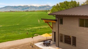 A modern house with a patio, a view of a green field, and mountains in the background. - Farmland Flat - Bozeman (Bozeman)