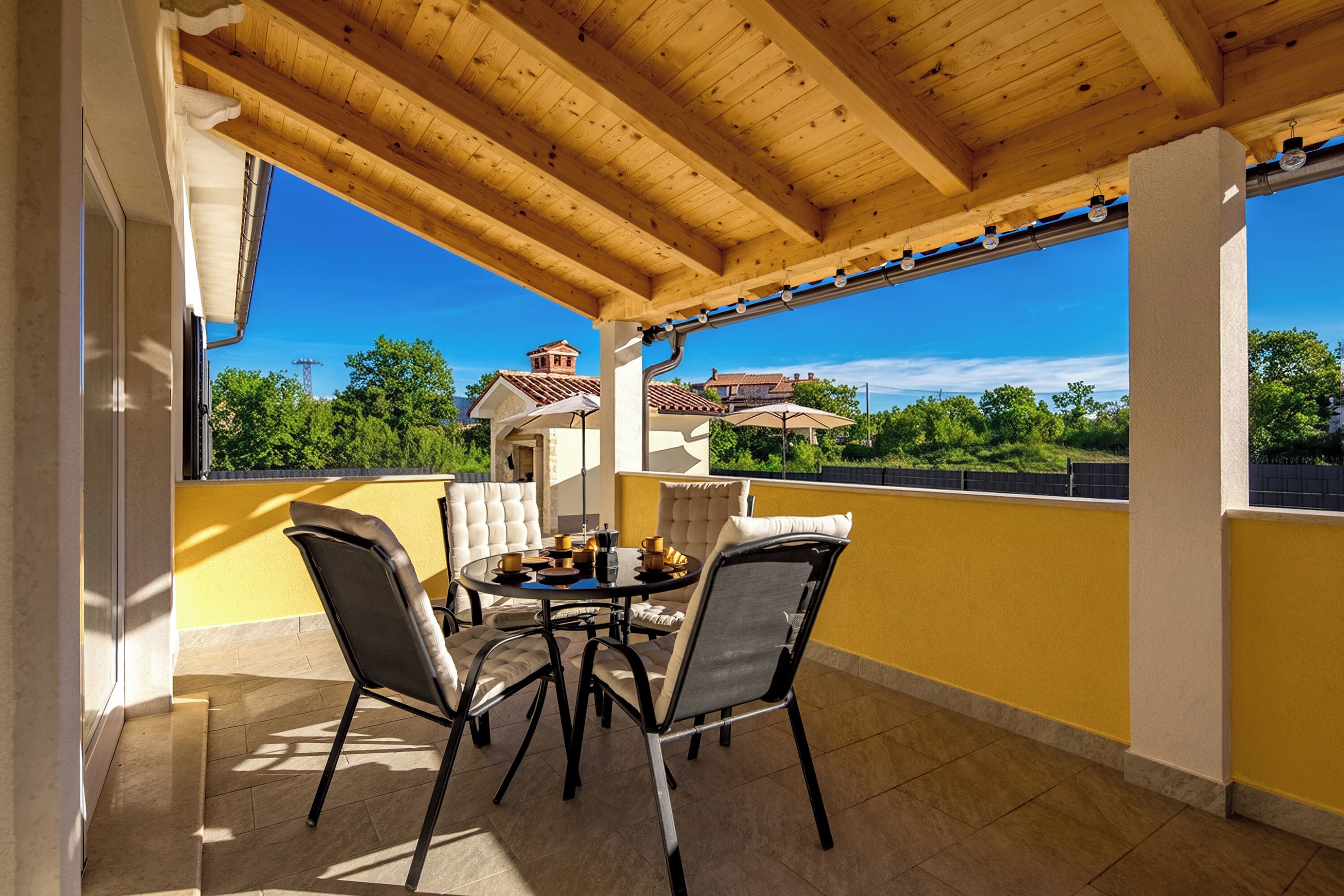 A covered patio with a wooden ceiling, a round table with chairs, and a view of a house and trees in the background.