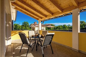 A covered patio with a wooden ceiling, a round table with chairs, and a view of a house and trees in the background. - Villa Berton – Heated Saltwater Pool (Sveta Nedelja)