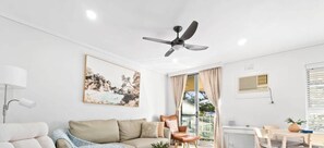 Una sala de estar moderna con un sofá, una silla, un ventilador de techo, un cuadro, una ventana con cortinas y una pequeña mesa con una planta.