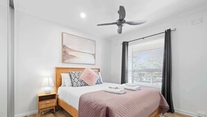 Ein Schlafzimmer mit Bett, Nachttisch, Lampe, Deckenventilator, Fenster mit Vorhängen und einem Bild an der Wand.