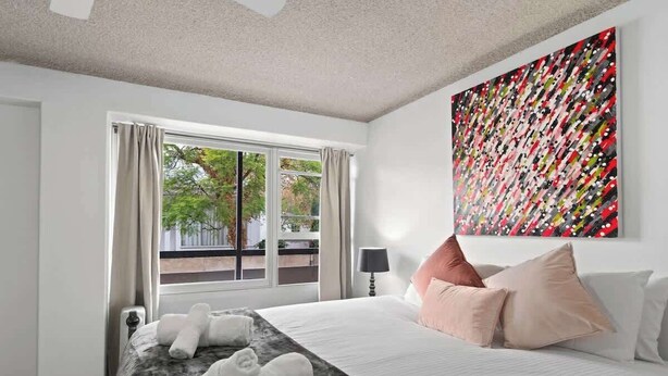 A hotel room with a bed, a window with curtains, a painting on the wall, and a view of trees outside. - North Adelaide View | 3BR & Free Parking (North Adelaide)
