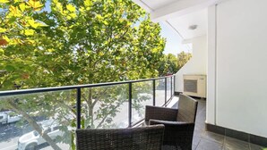 A balcony with wicker chairs, a glass railing, and a view of trees and a building. - Cozy 2BR Apt CBD Wi-Fi Free Parking (Adelaide)