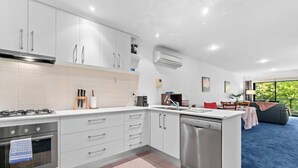 A modern kitchen with white cabinetry, a gas stove, an oven, and a dishwasher. There is a dining area with a table and chairs, and a large window providing a view of greenery outside. - Cozy 2BR Apt CBD Wi-Fi Free Parking (Adelaide)