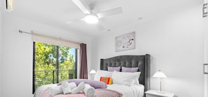 Un dormitorio con cama, ventilador de techo, ventana con cortinas, lámpara de noche y una obra de arte colgada en la pared.
