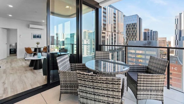 A modern apartment with a balcony, furnished with wicker chairs and a glass table, overlooking a cityscape. - CBD VTA Apartment | Free Parking | Walk Everywhere (Adelaide)