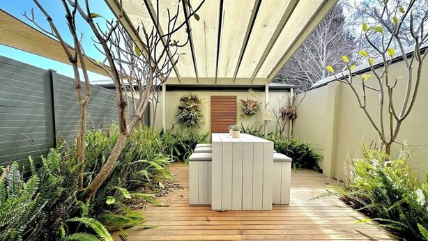Une terrasse en bois avec un espace salon extérieur blanc, entourée de verdure et agrémentée d’une pergola.