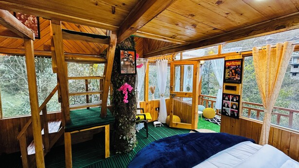 Eine Holzhütte mit einem Bett, einem kleinen Tisch und einem Stuhl. Mehrere Fenster bieten Blick auf Bäume.
