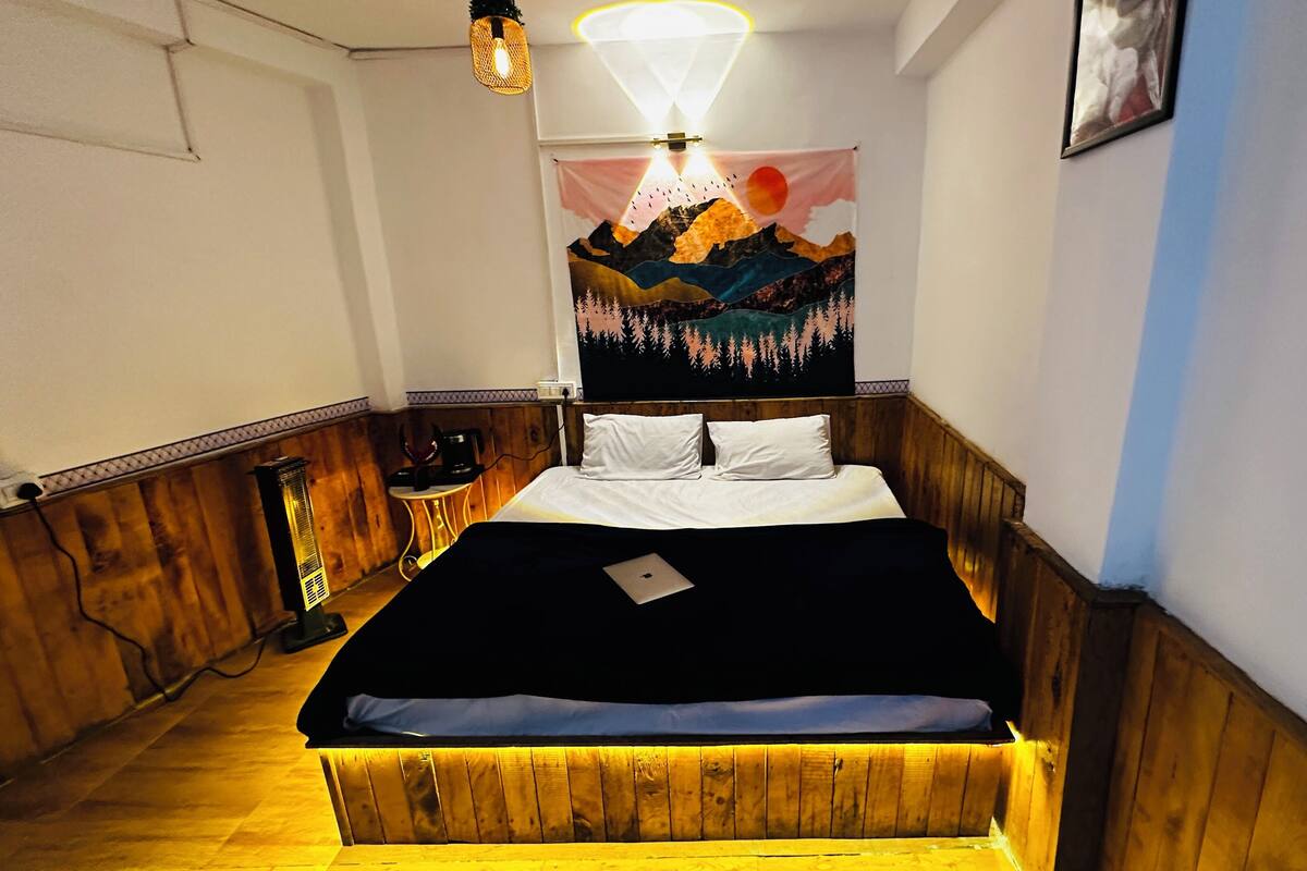 A bedroom with a wooden headboard, a bed with white bedding, a laptop on the bed, a wall tapestry, and a nightstand with a lamp.