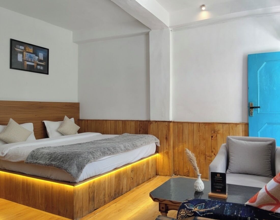 A modern hotel room with a wooden headboard, a bed with a gray blanket, a small table with a vase, and a blue door.