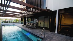 An indoor swimming pool with a sunroof, lounge chairs, and a glass door leading to a balcony. - High Street Apartments Toowong (brisbane)