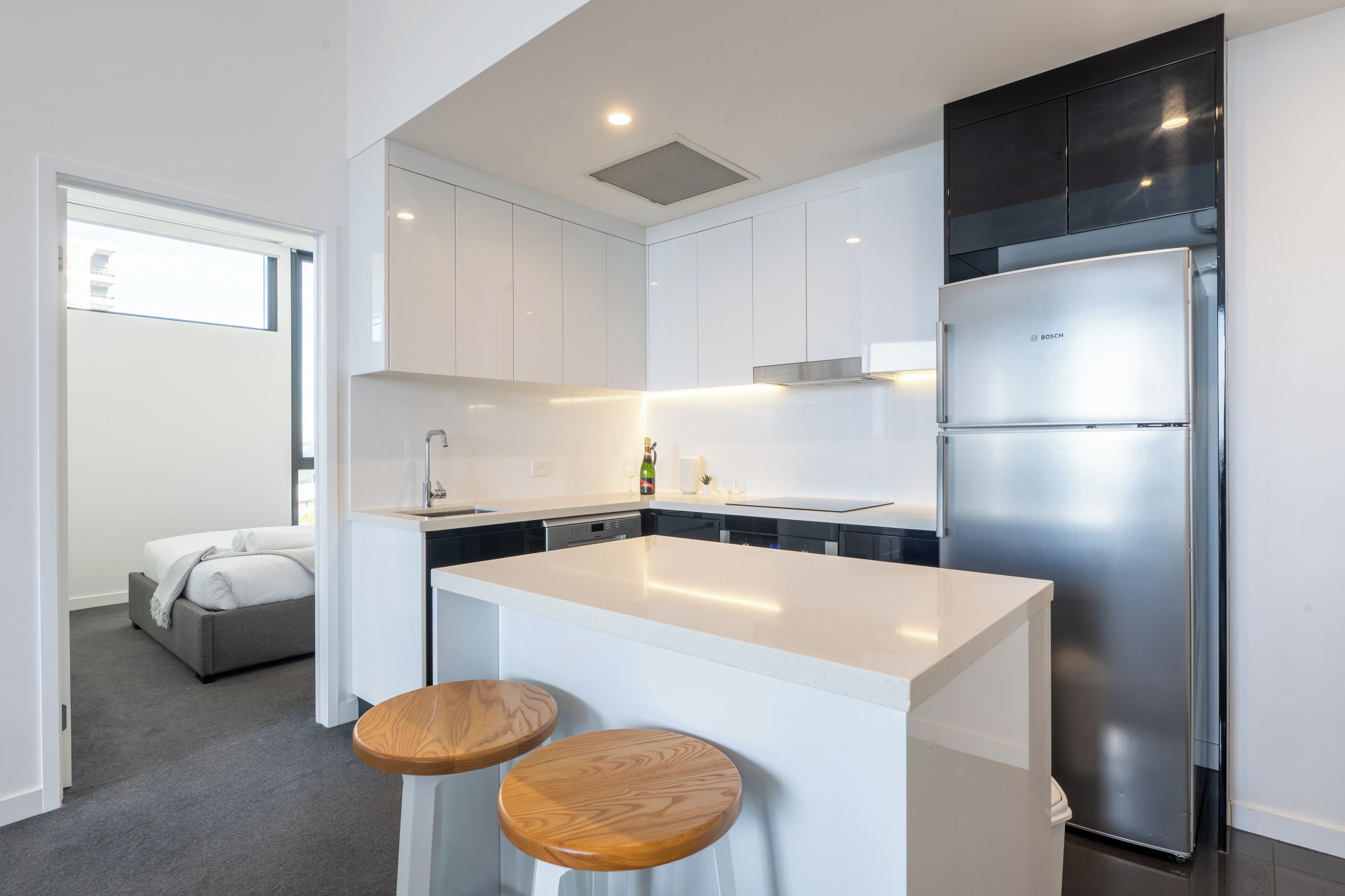 A modern kitchen with white cabinetry, a stainless steel refrigerator, and two wooden stools.