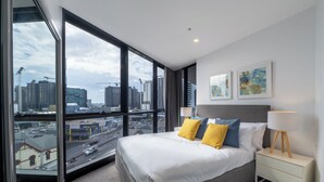 A hotel room with a large window, a bed, a bedside table, and two wall-mounted paintings. - High Street Apartments Toowong (brisbane)