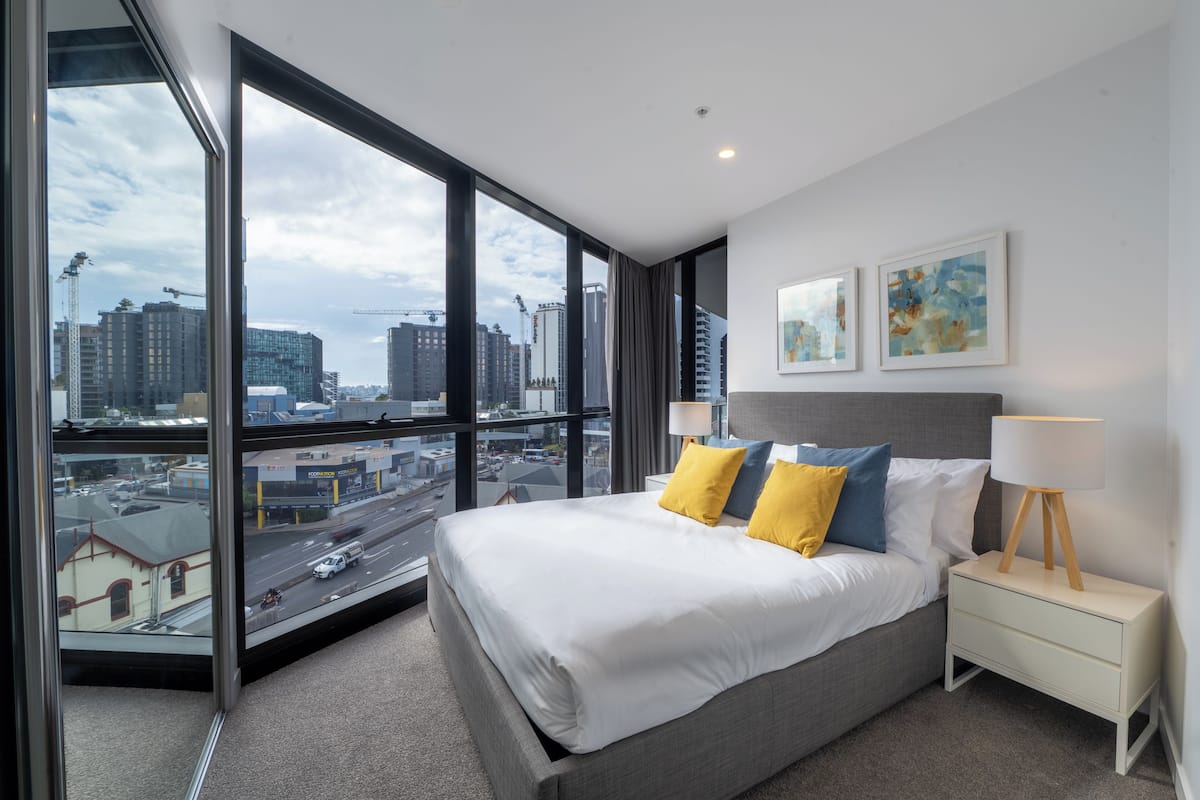 A hotel room with a large window, a bed, a bedside table, and two wall-mounted paintings.