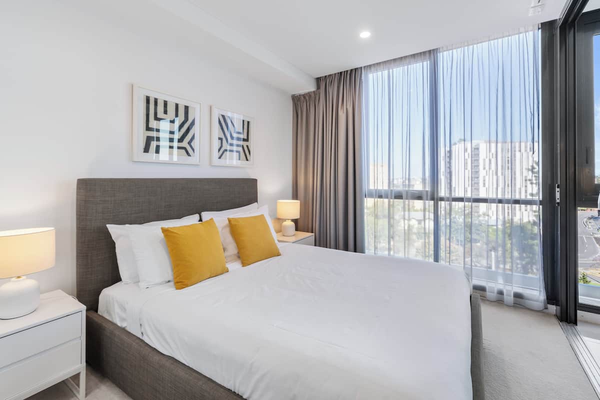 A modern hotel room with a large bed, two bedside tables with lamps, and a view of the city through floor-to-ceiling windows.