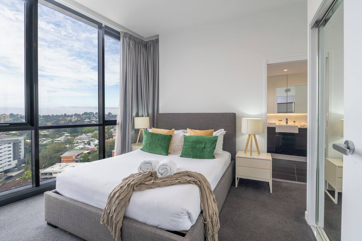 A modern hotel room with a large bed, a view of the city, a bathroom with a mirror, and a small bedside table.