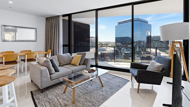 A modern living room with a sofa, coffee table, and chairs, large windows with a city view, and a painting on the wall. - High Street Apartments Toowong (brisbane)
