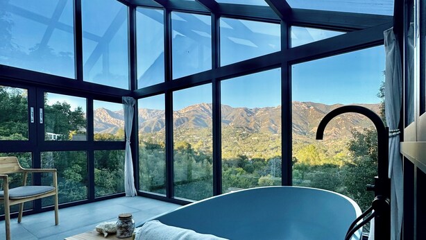 A modern bathroom with a large bathtub, a wooden shelf with a towel, and a view of mountains through large windows.