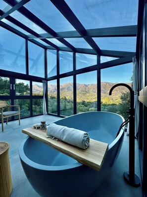 A modern bathroom with a large bathtub, a wooden shelf with a towel, and a view of mountains through large windows. - 3-Bdr Riviera Luxury Retreat - Designer Interiors, Heated Pool & Mountain Views (Santa Barbara)