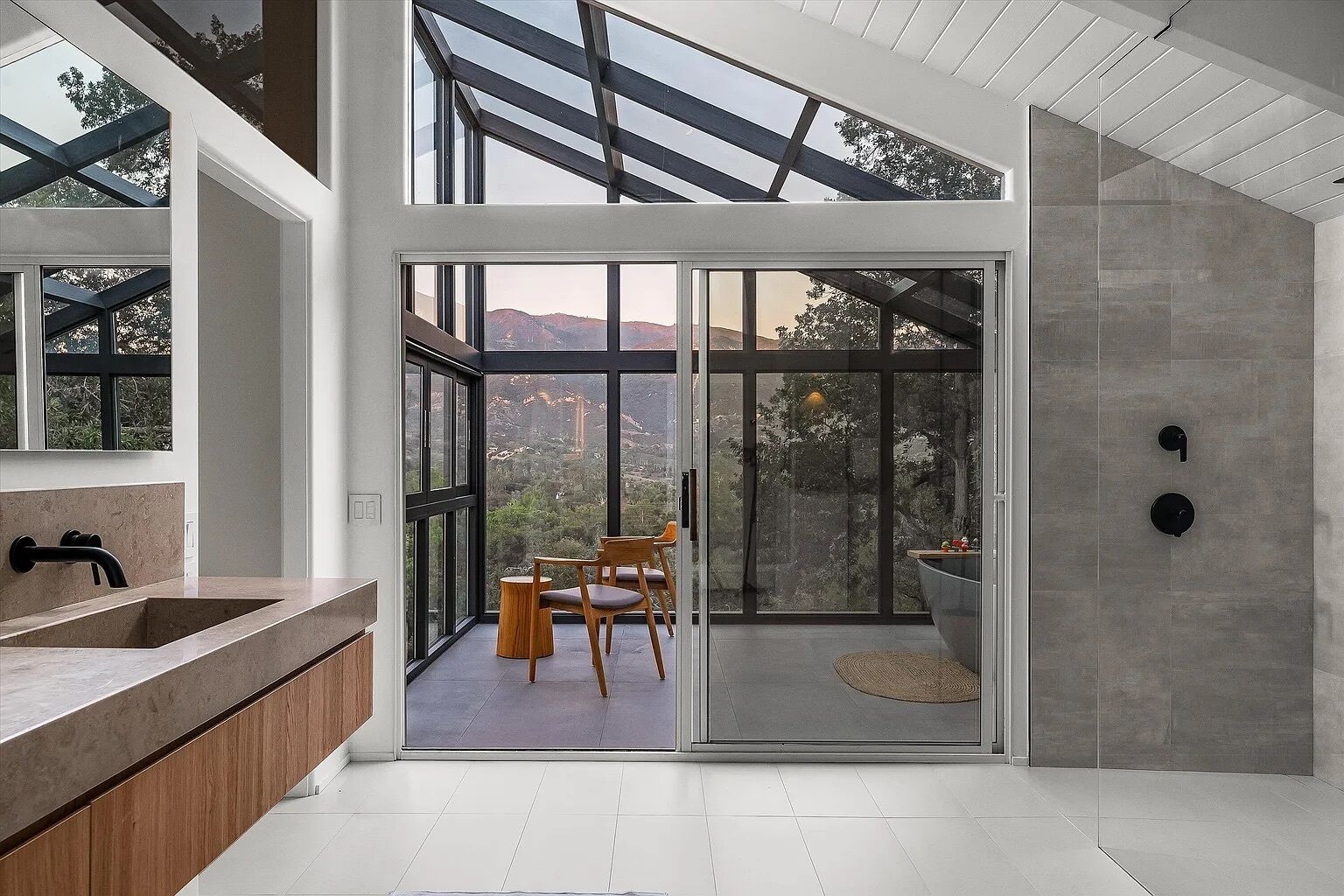 A modern bathroom with a large glass door leading to a balcony, a wooden vanity, and a view of the mountains.