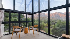 A room with large windows offering a view of a mountainous landscape. There are two wooden chairs and a small round table.
