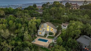 A modern house with a swimming pool surrounded by dense trees, overlooking a coastal area.