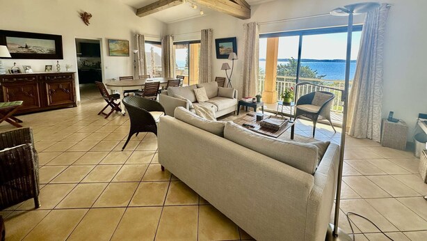 Ampla sala de estar com um sofá grande, mesa de jantar e cadeiras, varanda com vista para o mar e vigas de madeira aparentes no teto.