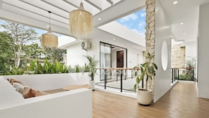 A spacious, modern interior with a wooden floor, white walls, and a stone accent wall. There are potted plants and a hanging light fixture. - Sadin Villa by Kozystay - Sanur (Kerobokan)