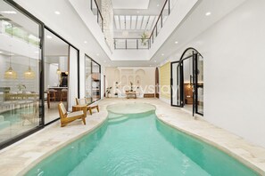 Et moderne poolområde indendørs med væg i glas, træloungestole og loft med indbygningsbelysning.