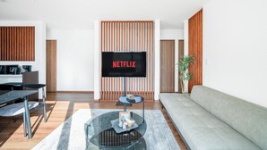 Ein modernes Wohnzimmer mit Flachbildfernseher an einer Holzwand, einem runden Glastisch, einem grauen Sofa und einem Essbereich mit Tisch und Stühlen.