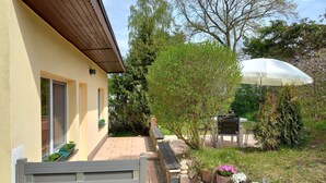 Eine Terrasse mit einer Holzbank, einem weißen Sonnenschirm und einem Garten mit verschiedenen Pflanzen und Blumen.