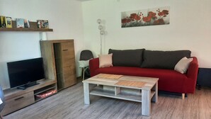 Ein Wohnzimmer mit einem roten Sofa, einem Holztisch, einem Fernseher auf einem Holzschrank und einem Bücherregal mit verschiedenen Gegenständen.