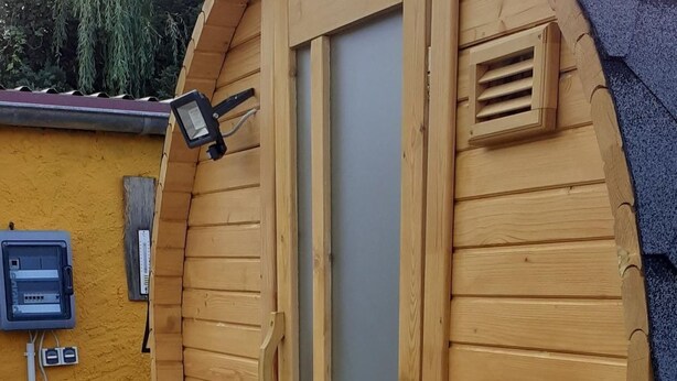 Eine Holzhütte mit großem Fenster und T ür, einem kleinen Lüftungsgitter und einer Außenleuchte an der Außenwand.