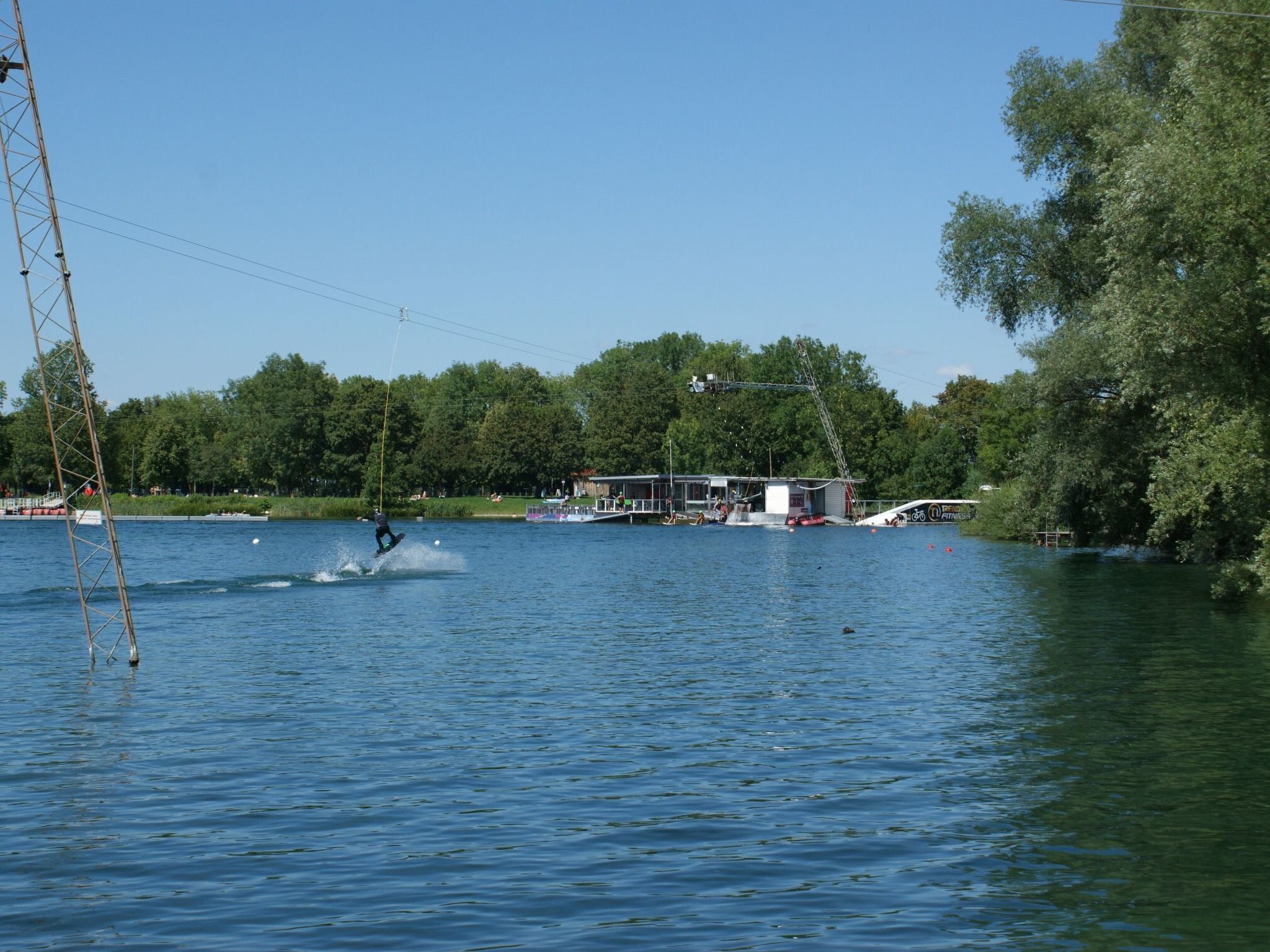 Escena junto al lago con un practicante de wakeboard, un muelle y un edificio con porche cubierto.