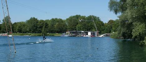 Escena junto al lago con un practicante de wakeboard, un muelle y un edificio con porche cubierto.