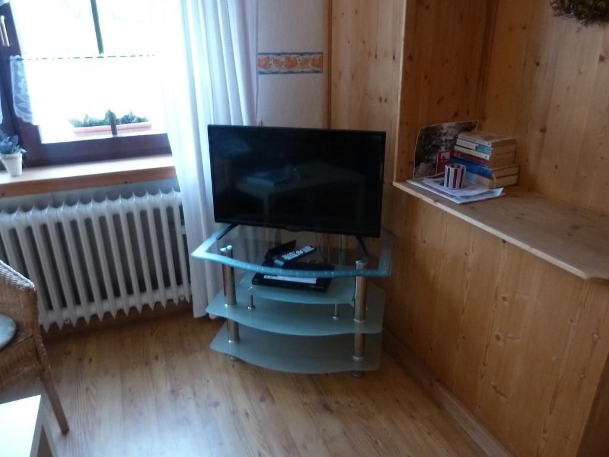 Ein Wohnzimmer mit Fernseher auf einem Glastisch, Heizkörper, Holzböden und einem Fenster mit Vorhängen.