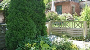 Een tuin met een hoge naaldboom, een houten poort en een bakstenen gebouw met groene luiken.
